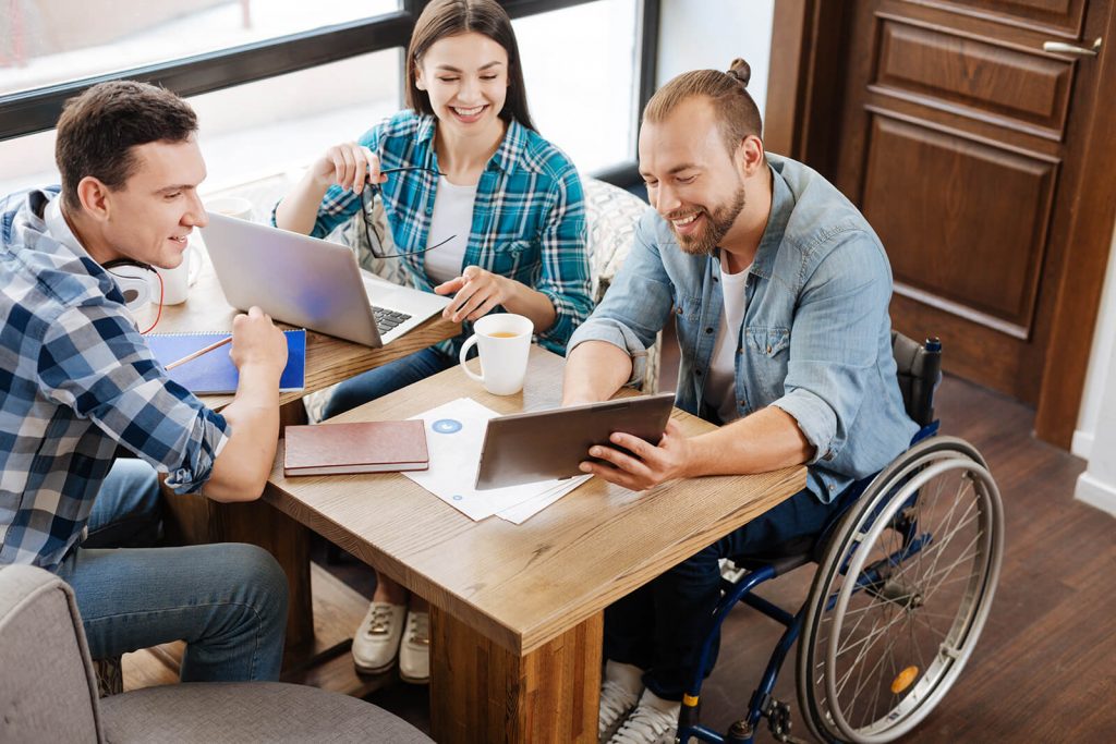 Drei Junge Menschen sitzen am Tisch und schauen sich etwas auf einem Tablet an. Einer davon sitzt im Rollstuhl. SAB NRW Bochum Startseite